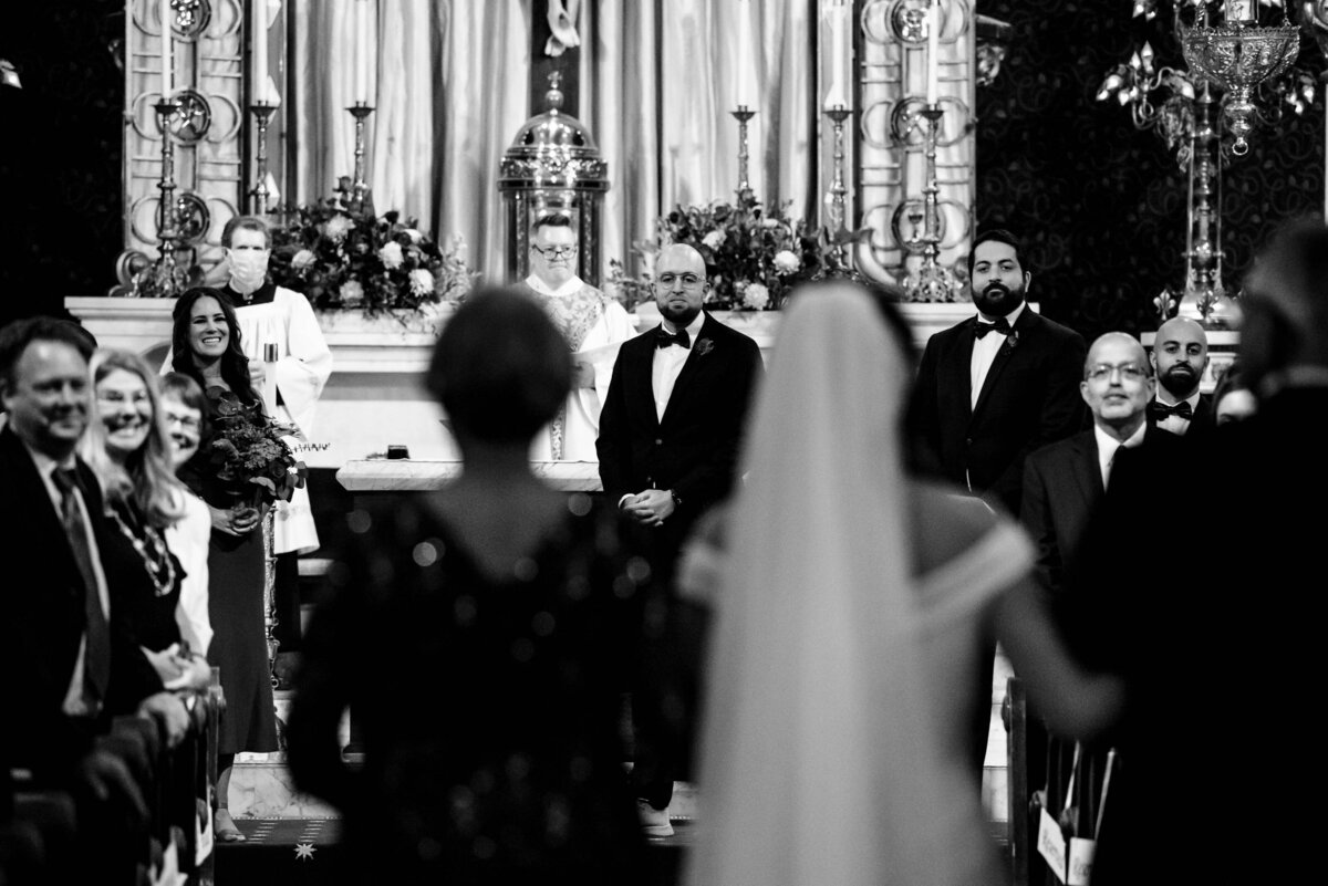 Black and white photograph of the bride walking down the aisle with her parents in the foreground at Basilica of the Sacred Heart of Jesus in Atlanta with the focus on the grooms face between the mother and daughter's shoulders