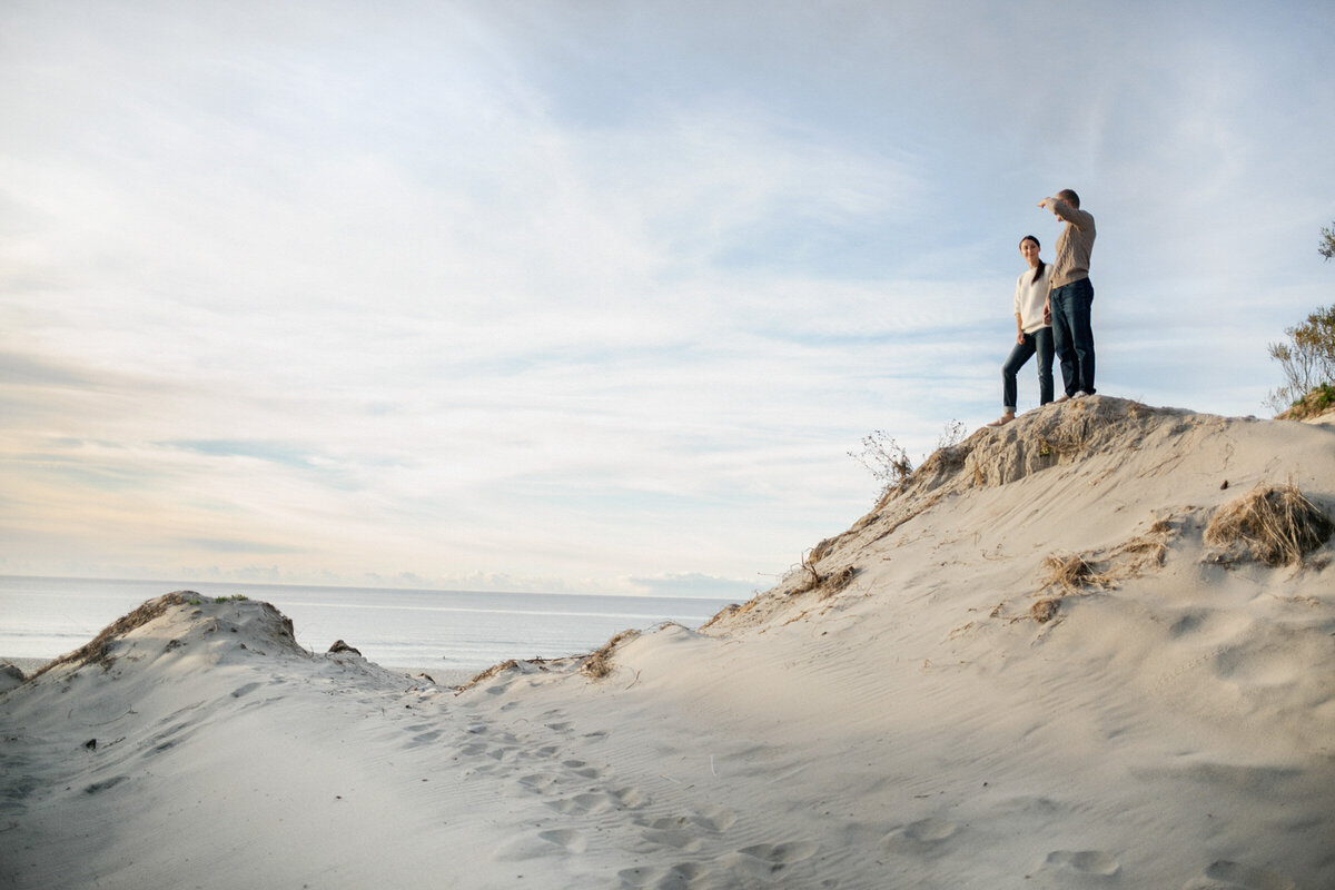 Couple standing on a tall dune by the ocean, looking out toward the horizon during their engagement session