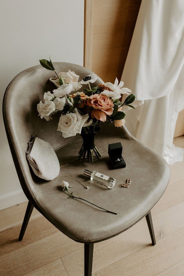 This stunning flat lay features a modern bridal bouquet in white, terracotta, black, and blush hues, resting elegantly on a chair. Surrounding the bouquet are the bride's ring, clutch, perfume, and jewelry, creating a beautiful, cohesive look. Ideal for couples in Ottawa seeking unique floral designs and personalized wedding details.