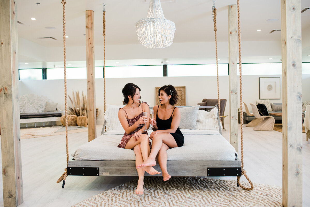 bride sitting on a bed in her bridal suit with her bridesmaid chatting as they get ready for this colorado wedding captured by denver wedding photographer