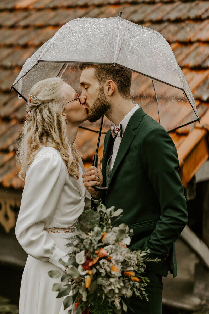 Mariés échangeant un bisou sous un parapluie transparent pendant un temps pluvieux devant un toit de tuiles rouges. Immortalisés par Laura Termeau, photographe professionnelle en Vendée.