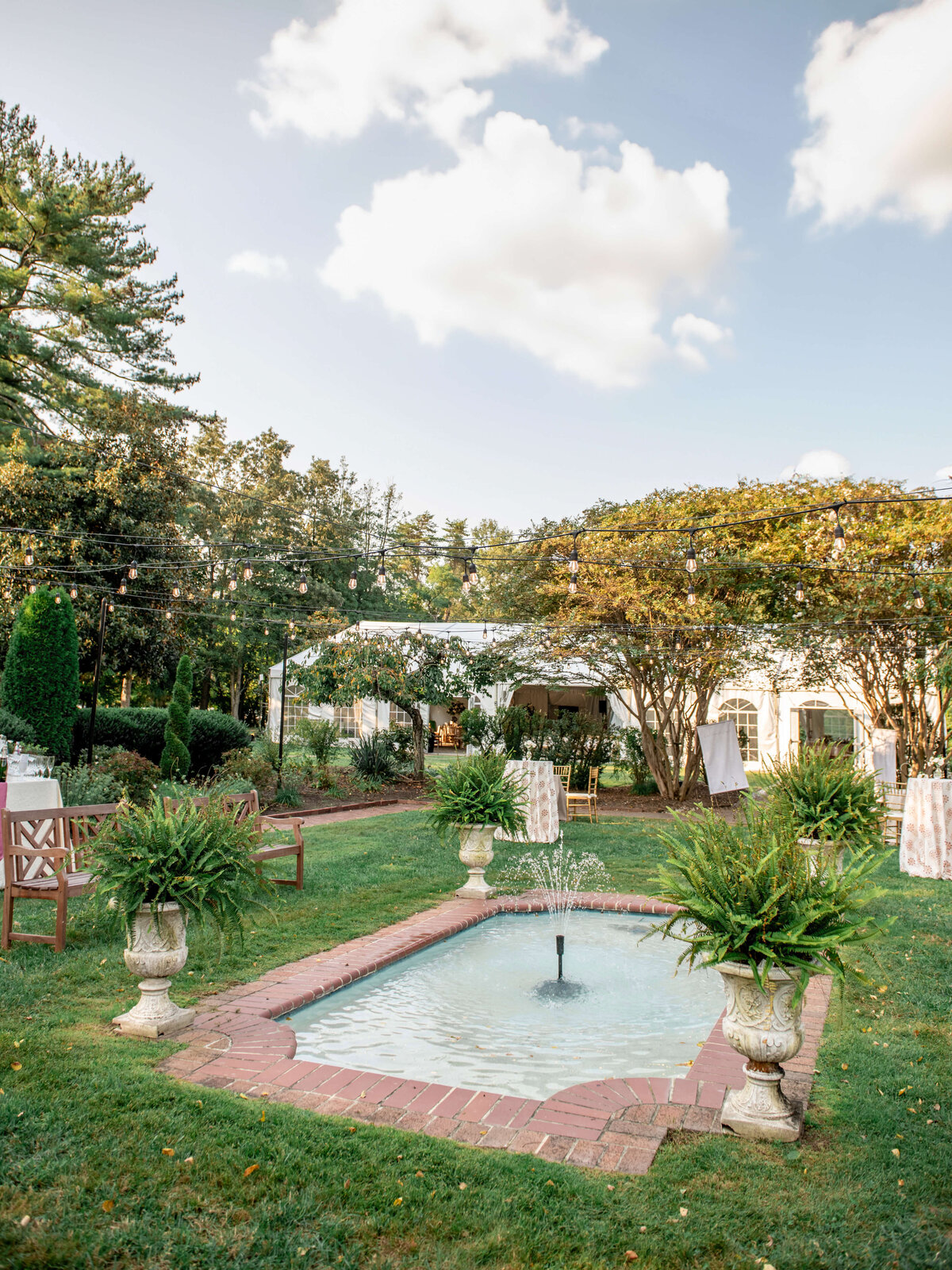 A tranquil garden scene features a small fountain surrounded by potted plants and a brick pathway. Benches are placed nearby, and string lights are hung above. Trees and a building form the backdrop under a partly cloudy sky.