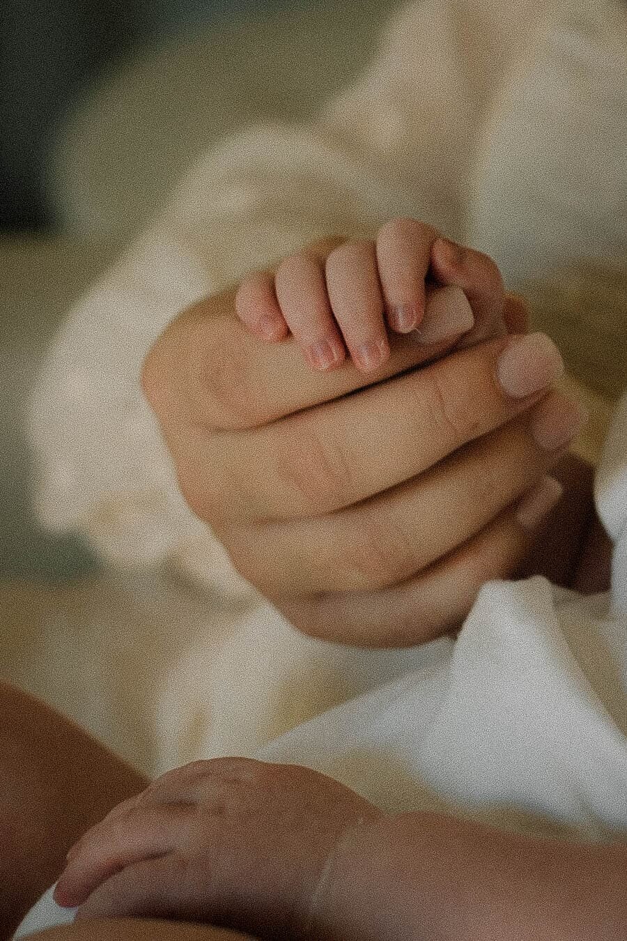 Lifestyle newborn photography detail of a baby’s tiny hand holding onto mama’s fingers, capturing tender connection at home.