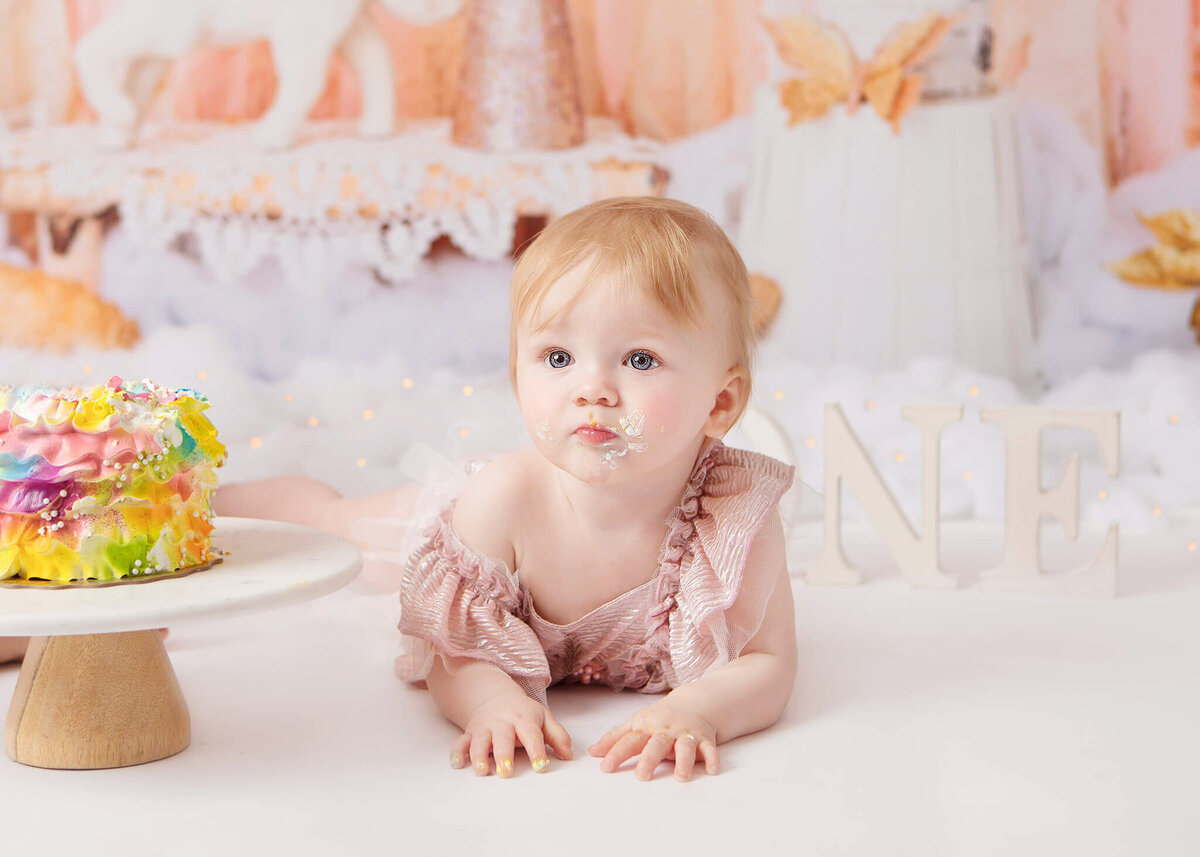 Baby leaning forward by her rainbow cake for her first birthday cake smash