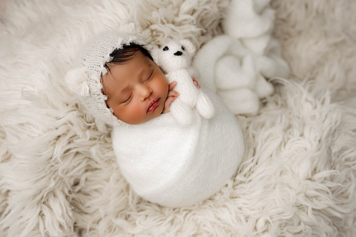 Newborn girl wrapped in a fuzzy white knit, asleep on a white shag blanket while cuddling a tiny white bear and wearing a bear-ear bonnet.