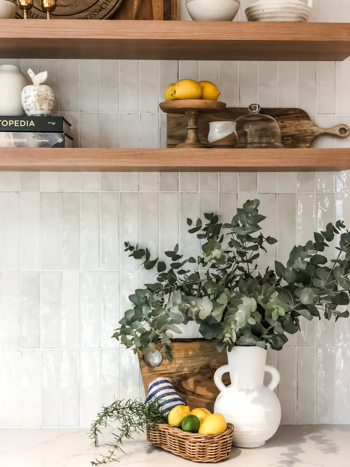 Kitchen shelves with lemons and decor, eucalyptus in a white vase, and a basket with fruit and herbs on the counter embody bespoke interior design Newcastle NSW.