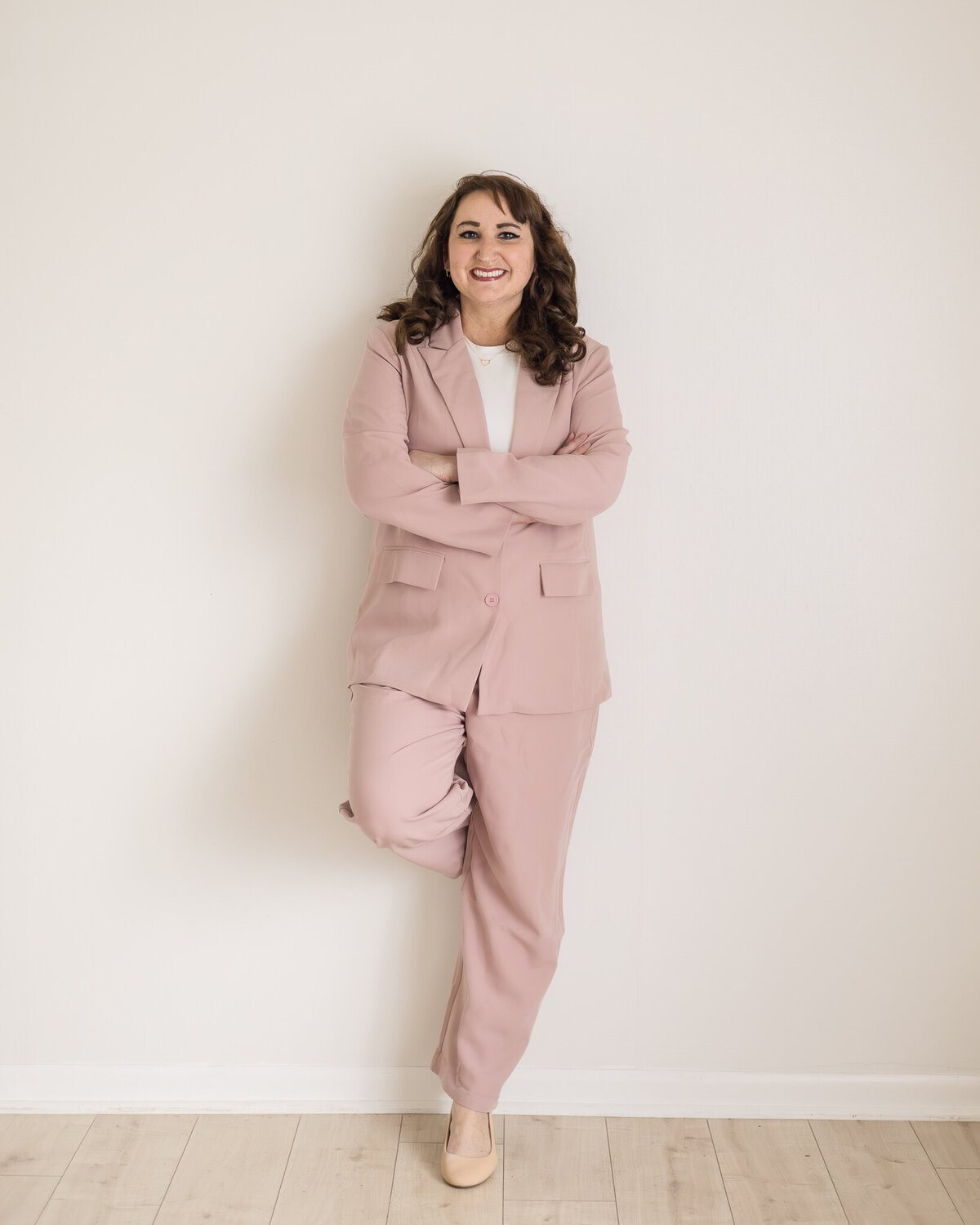 Dark brunette woman standing against a wall with one foot against the wall, wearing light pink pants and matching dress jacket with a white shirt under the jacket, and heels, smiling at the camera