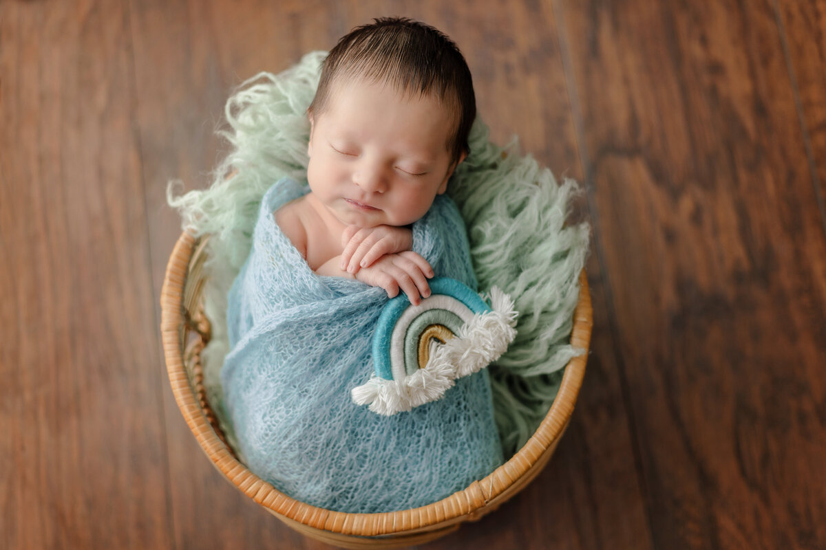 Newborn Photographer,  a baby is swaddled and sleeps in a basket holding a woven rainbow