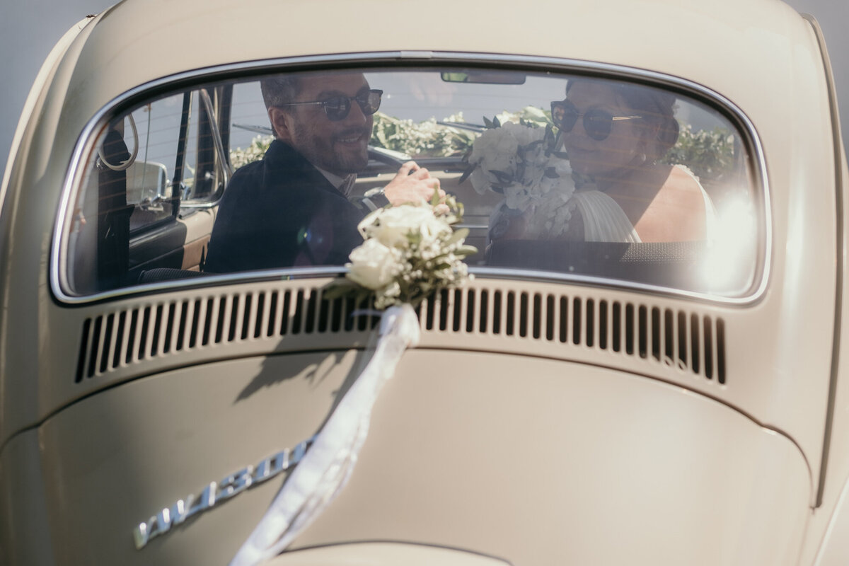 Von hinten sehen wir durch die Heckscheibe des Retro Hochzeitsautos auf das zurückblickende Hochzeitspaar.