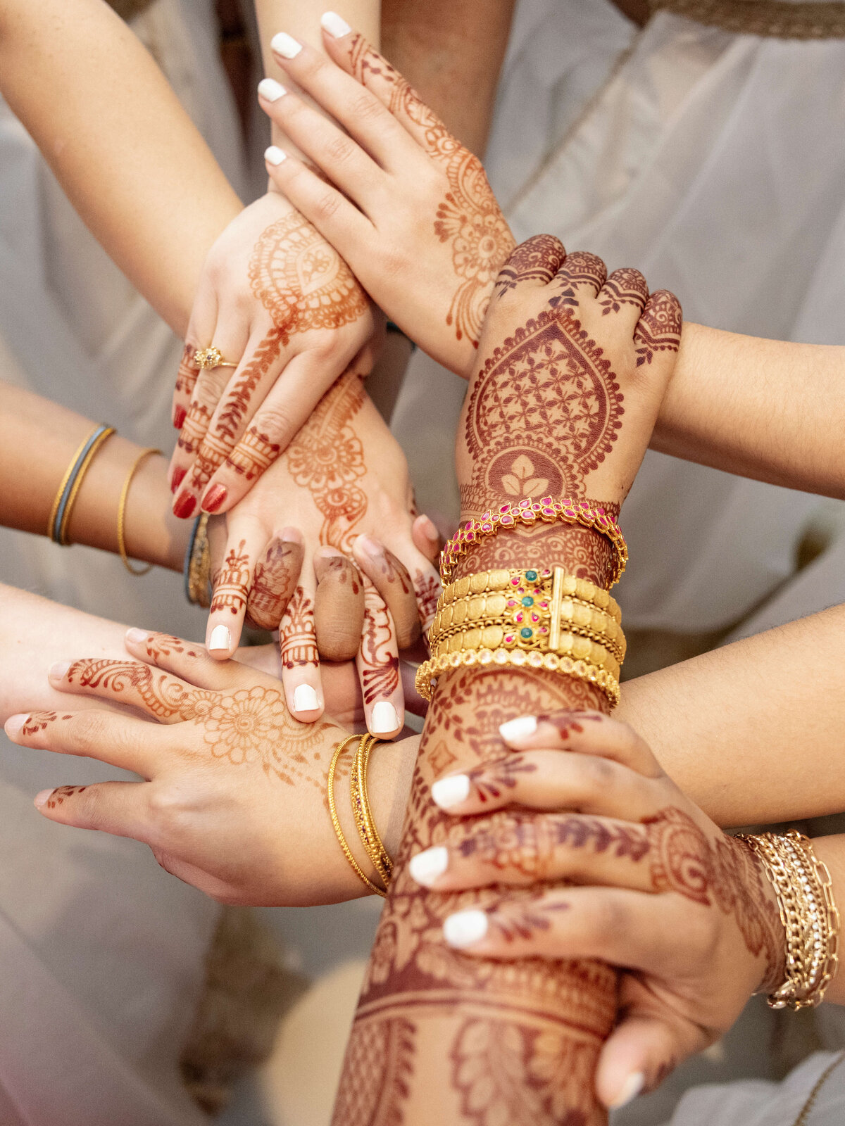 A group of hands adorned with intricate henna designs and gold bangles forming a circle. The hands vary in position, showcasing the detailed artistry of the henna patterns on the palms and fingers.