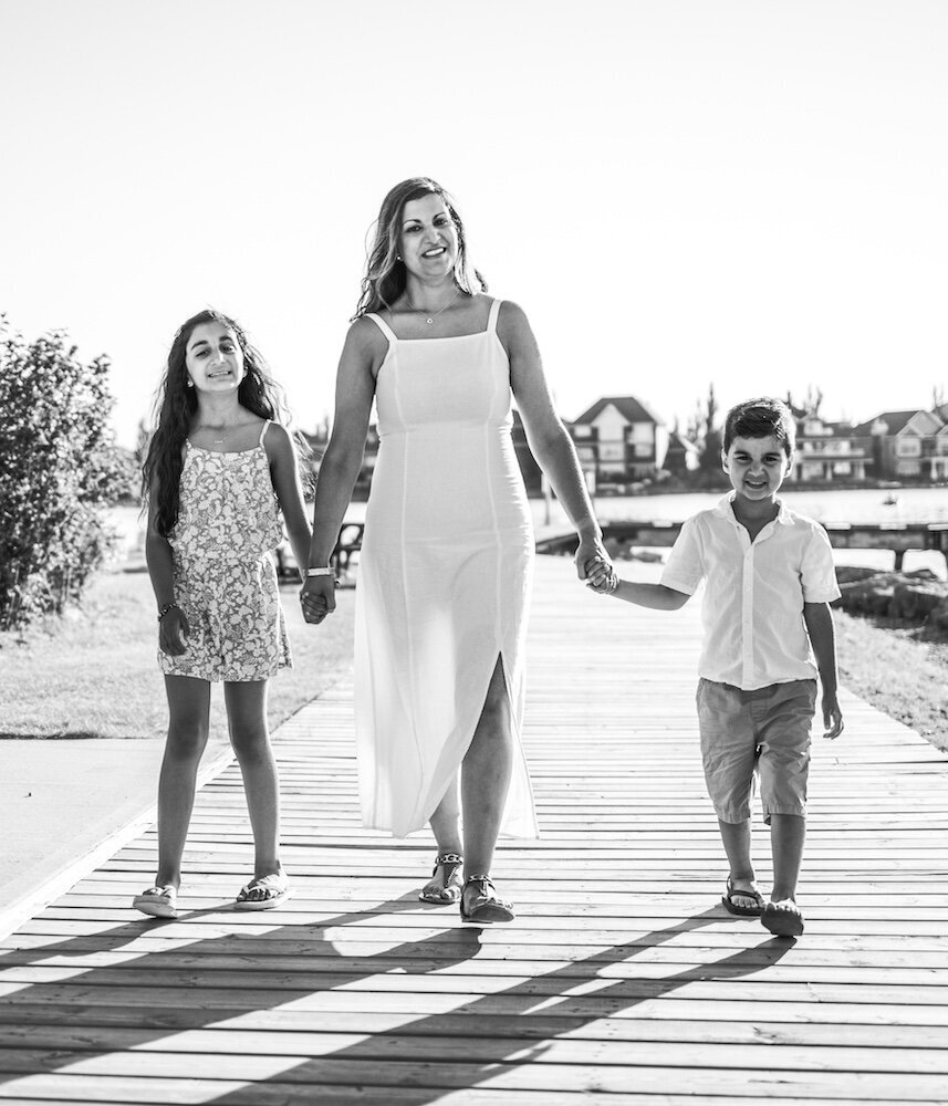 A mom in a white dress is walking near a lake holding her son and daughter's hands