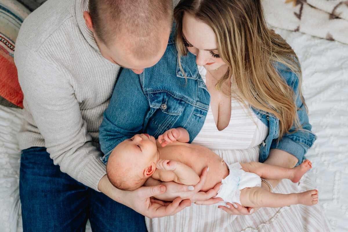 A top-down view of a couple holding their newborn baby. The mother, dressed in a striped dress and denim jacket, and the father, in a light sweater, lovingly gaze down at their baby, who is resting in their arms.