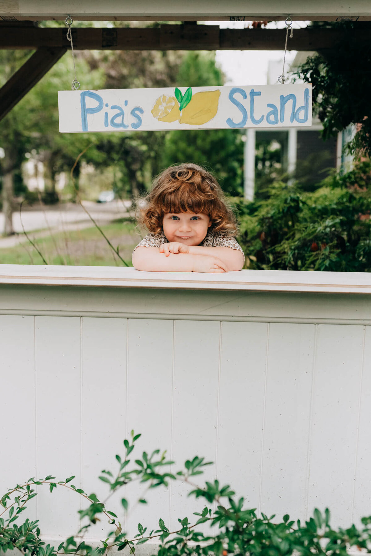 A toddler with red hair stands outside in her lemonade stand