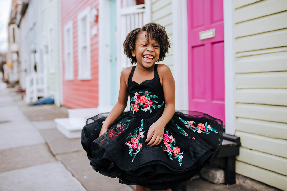 African American Girl wearing a black floral dress and laughing sitting on a white step near a pink door in Fells Point Baltimore Maryland