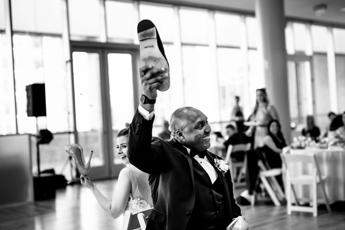 A joyful black and white photo of a man in a tuxedo raising a bottle of wine in celebration, with wedding guests blurred in the background.