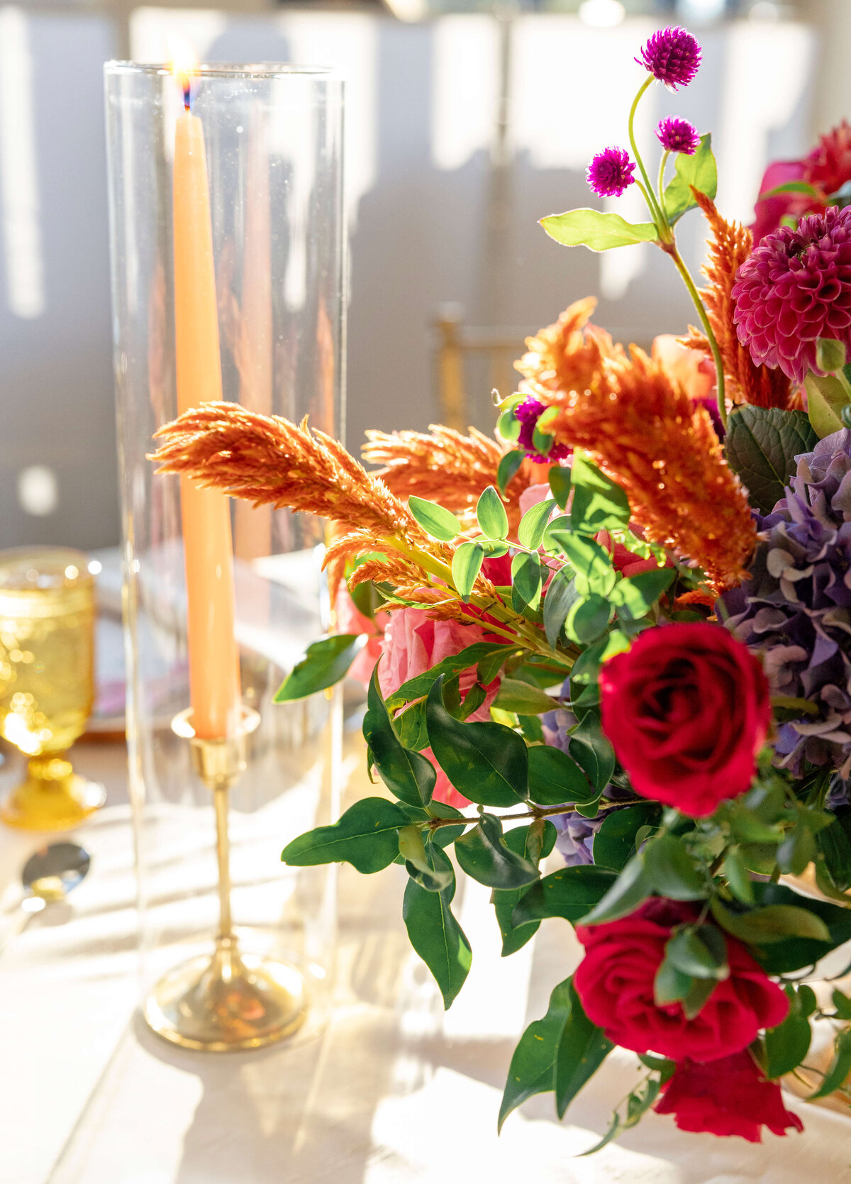 A vibrant floral arrangement with red roses, purple and pink flowers, and orange accents sits next to a tall lit candle in a glass holder on a sunlit table. Gold and glass decor pieces are visible in the background.