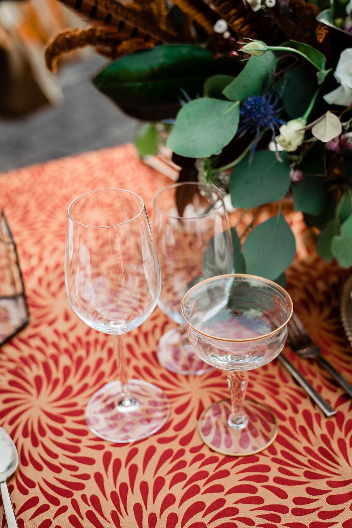 A stemmed red wine glass, a stemmed white wine glass and a gold rimmed coupe glass sit on an orange and rust textured table cloth next to a centerpiece of greenery at Rippa Villa.
