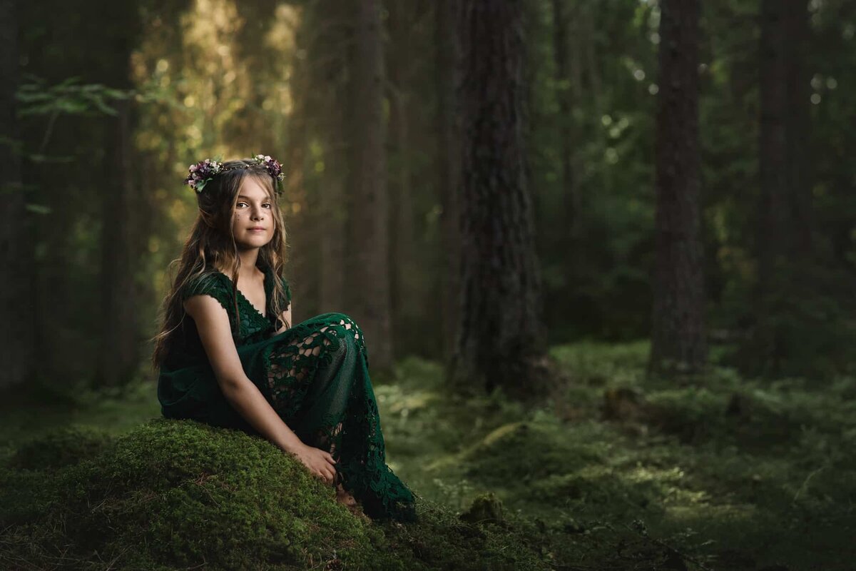 Sagolikt fotografi på flicka med alvöron och blomsterkrans i lila färger. Hon sitter på en mossig sten i skogen och solljuset glittrar bakom henne.