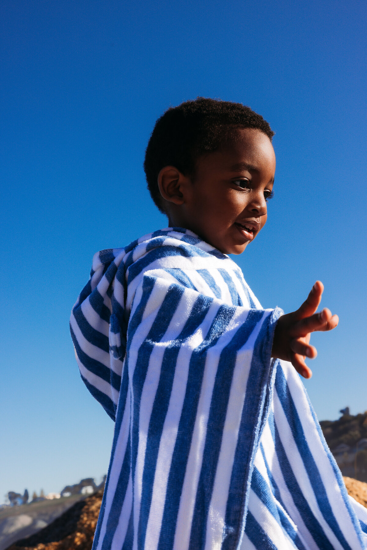 Smiling boy in a blue and white striped poncho on a sunny beach, photographed for Swim Essentials