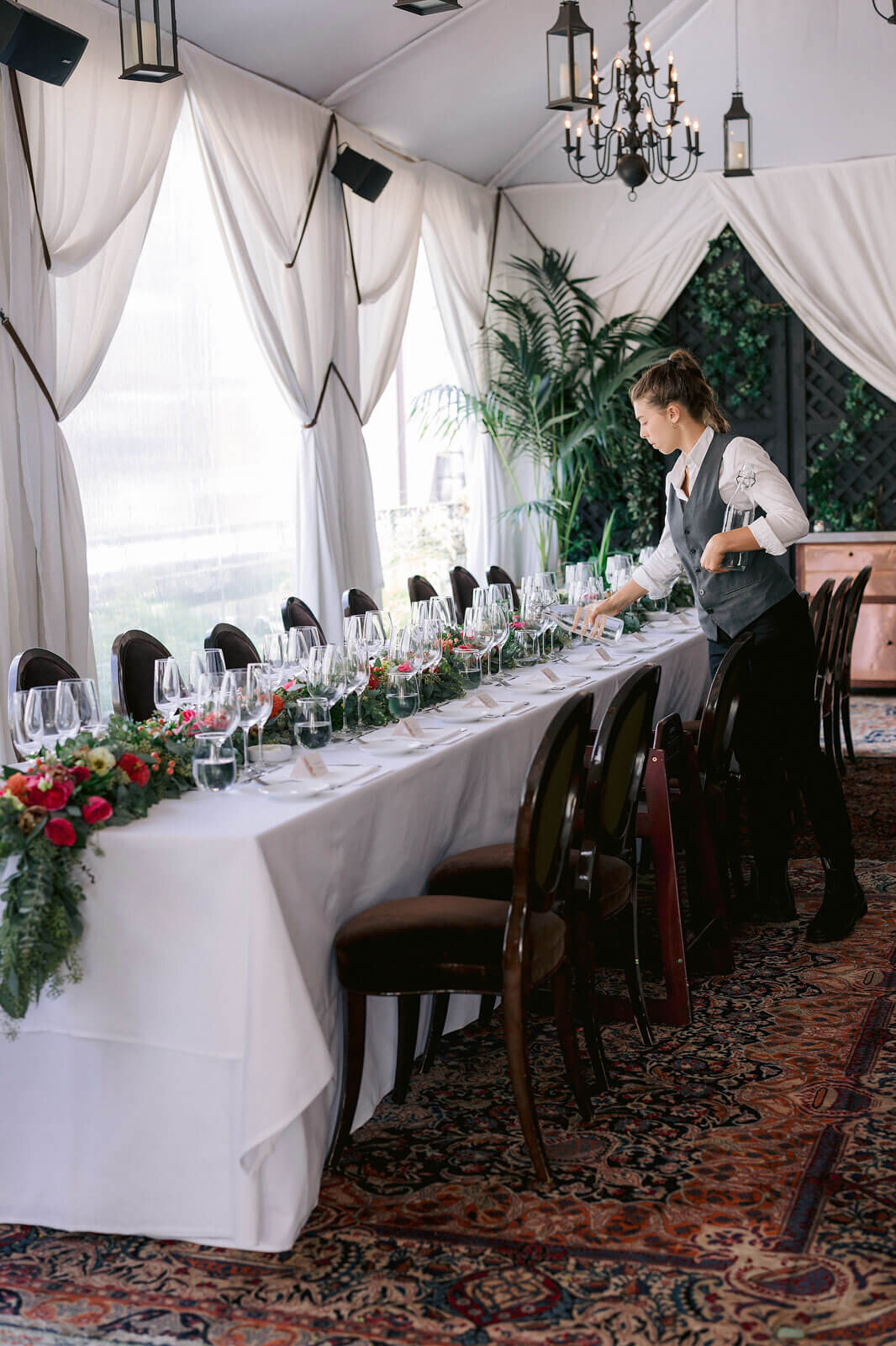 A crew is pouring water on wine glasses on the wedding dining table with red flower centerpieces. Image by Jenny Fu Studio