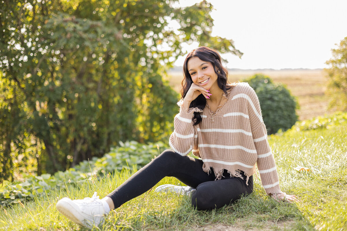 high school female senior, with long black hair, sitting in grass with a trees behind her, looking at the camera smiling, wearing black leggings, and light tan and white stripped shirt