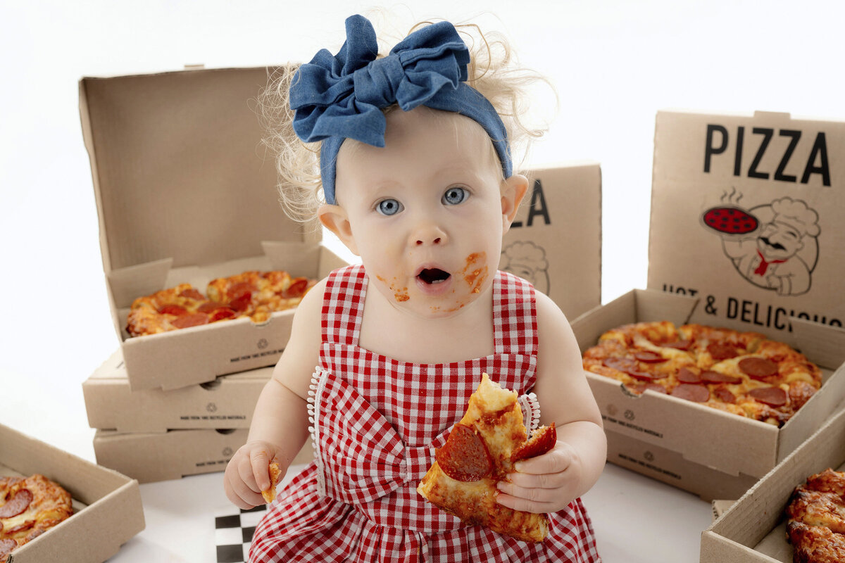 Toddler in a checkered outfit holding a slice of pizza, with pizza boxes surrounding them.
