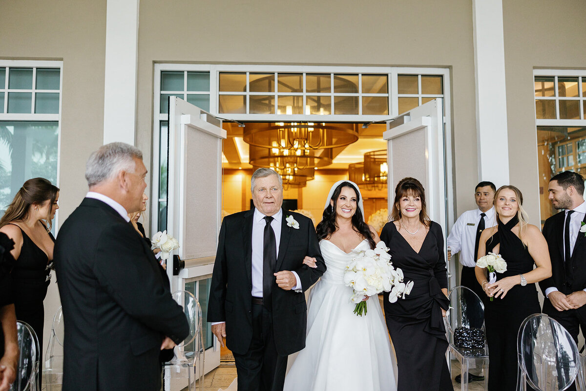 The bride walking into the ceremony with her parents by her side, radiating elegance and joy during Ashley and Cameron's micro wedding at Ocean Reef in Key Largo, captured by Claudia Amalia Photography.