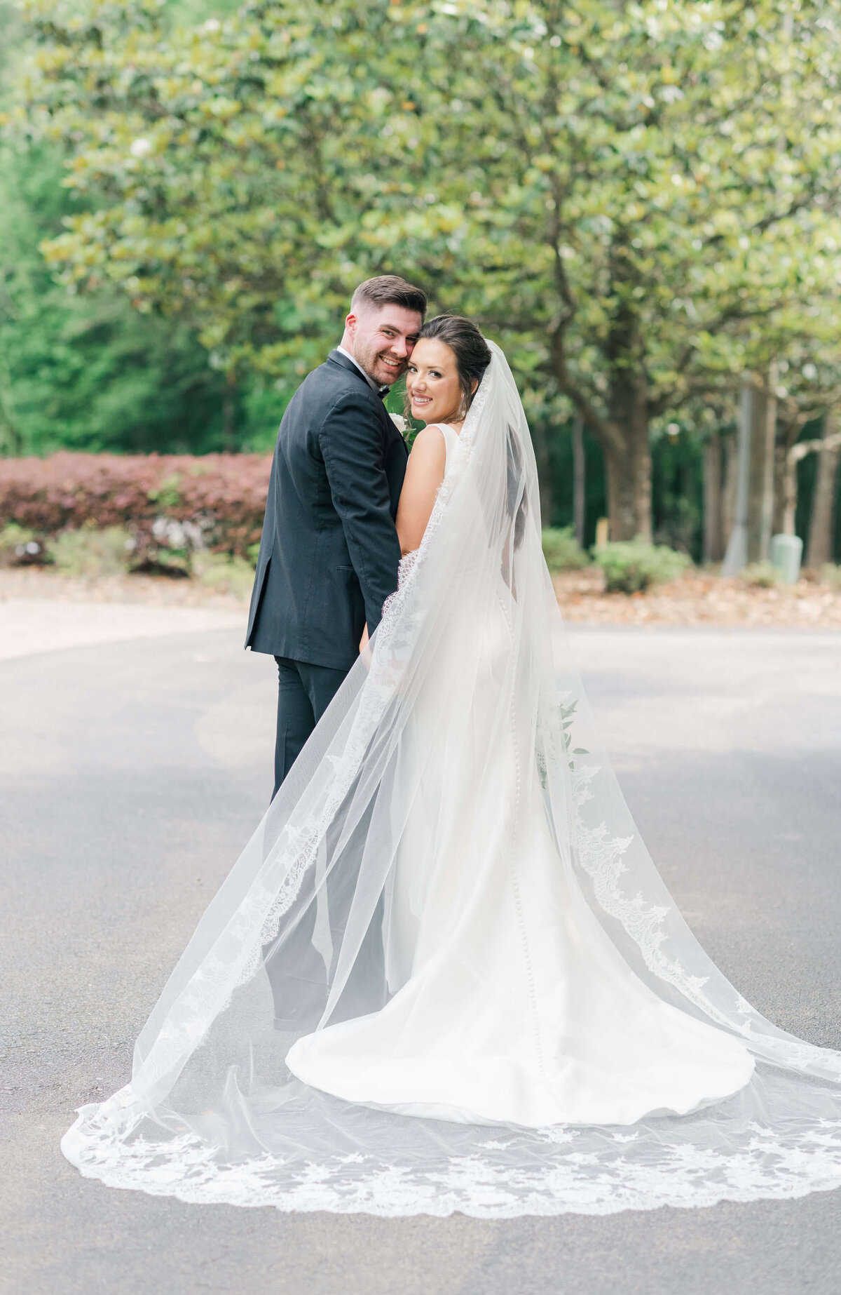 Bride and groom standing together with backs toward camera and heads turned smiling at camera while brides veil is sprawled out to see details