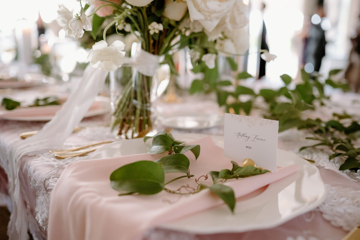 Wedding day stationery featuring elegant place card with guest name, blush napkin, and greenery detail.