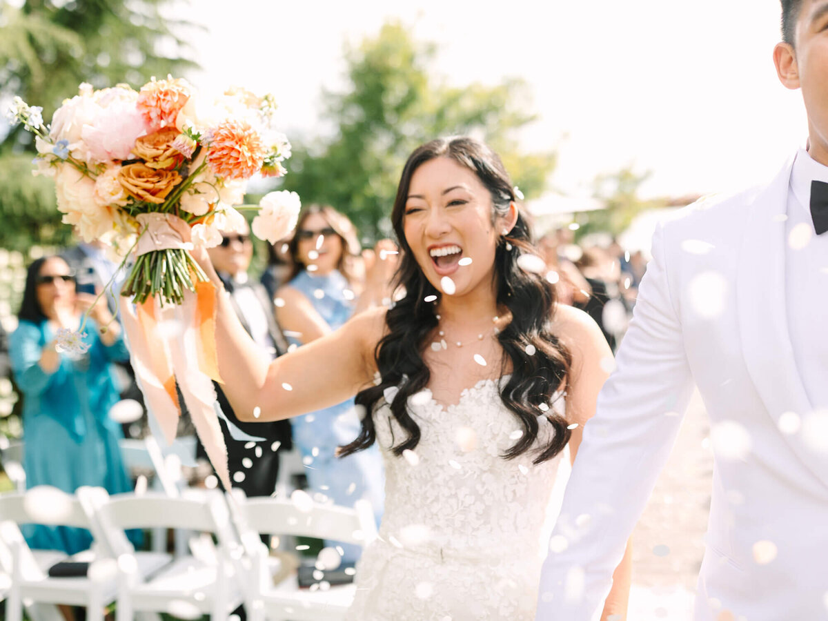 A bride in a lace gown joyfully holds a vibrant bouquet while walking down the aisle, surrounded by guests and confetti.