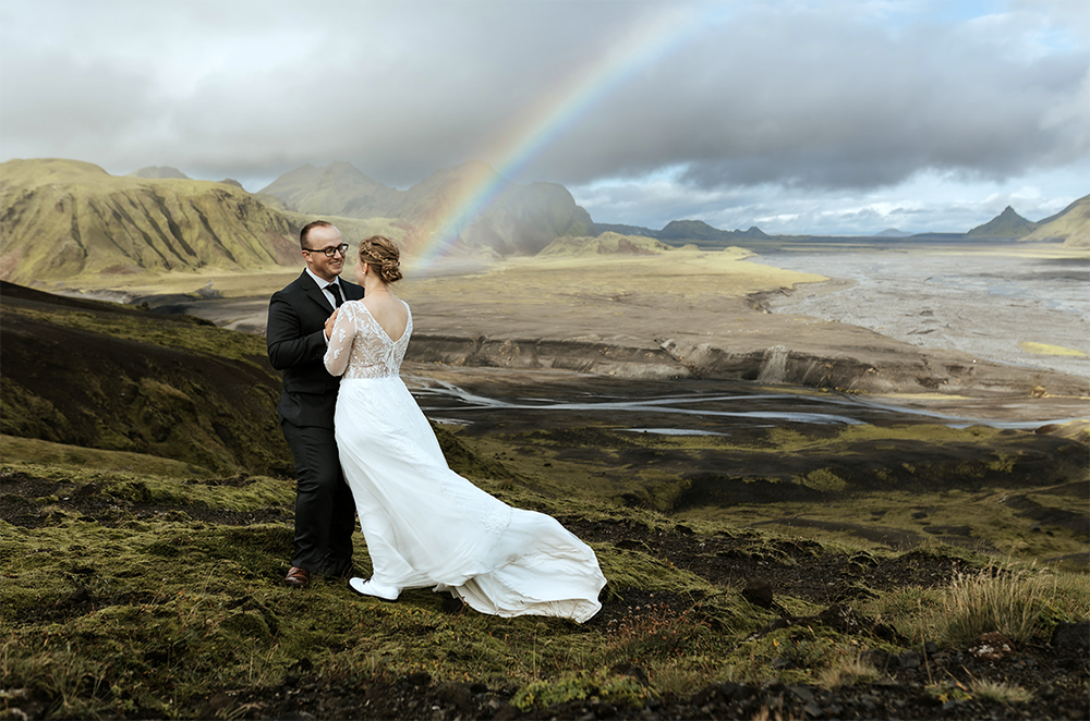 married couple is hugging in the middle of the highlands with mountains in the background and a rainbow on their wedding day in Iceland