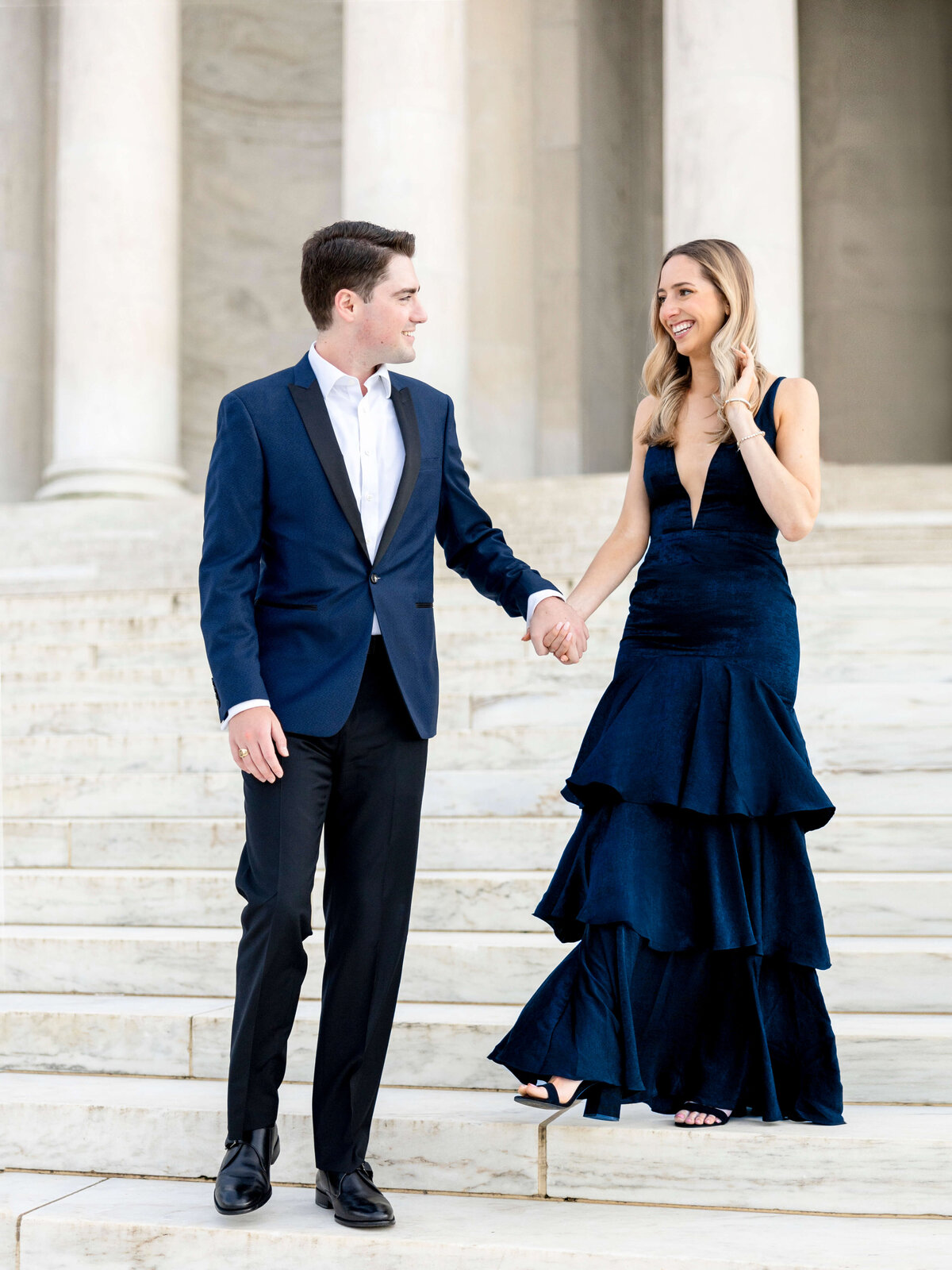 A couple dressed elegantly, standing on stone steps. The man wears a blue suit, and the woman is in a deep blue tiered gown. They hold hands, smiling at each other, in front of a building with tall white columns.