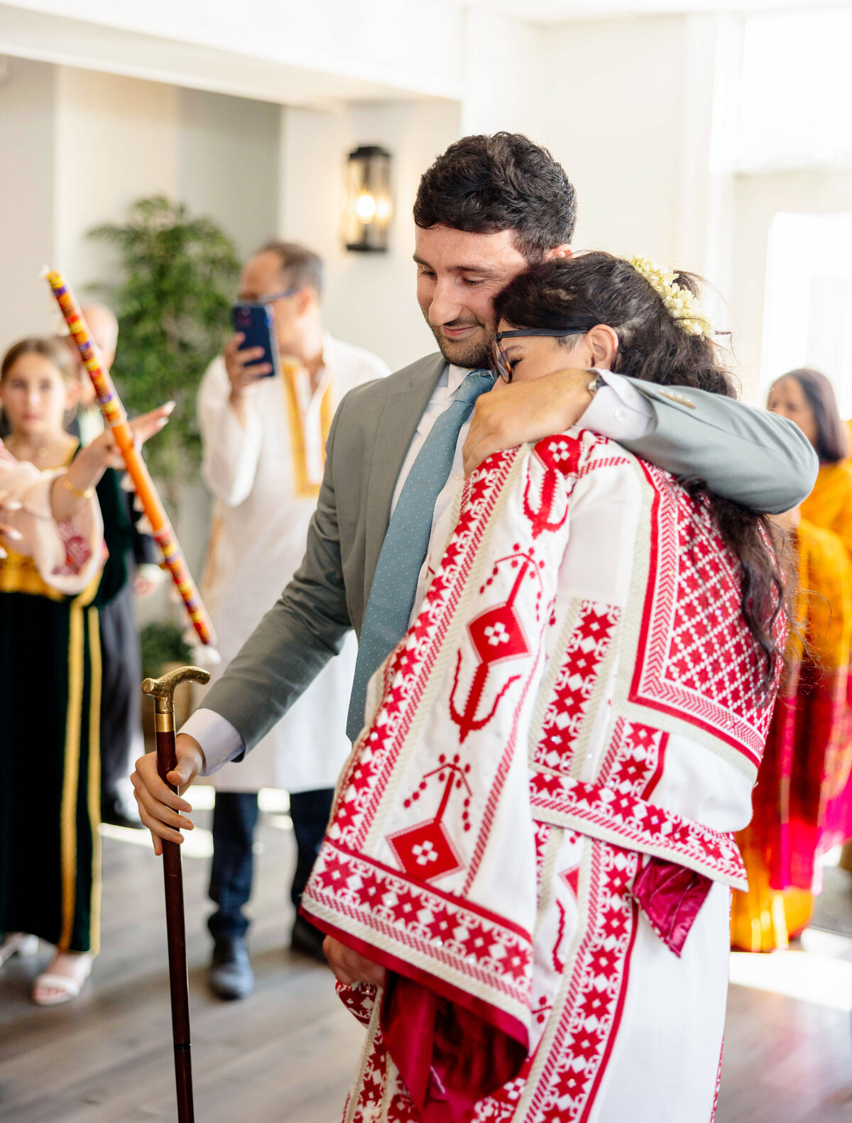 A man in a suit and a woman in a traditional red and white dress hug warmly at an indoor gathering. A cane is in the mans hand. People in the background wear festive clothing, and one person is taking a photo with a phone.