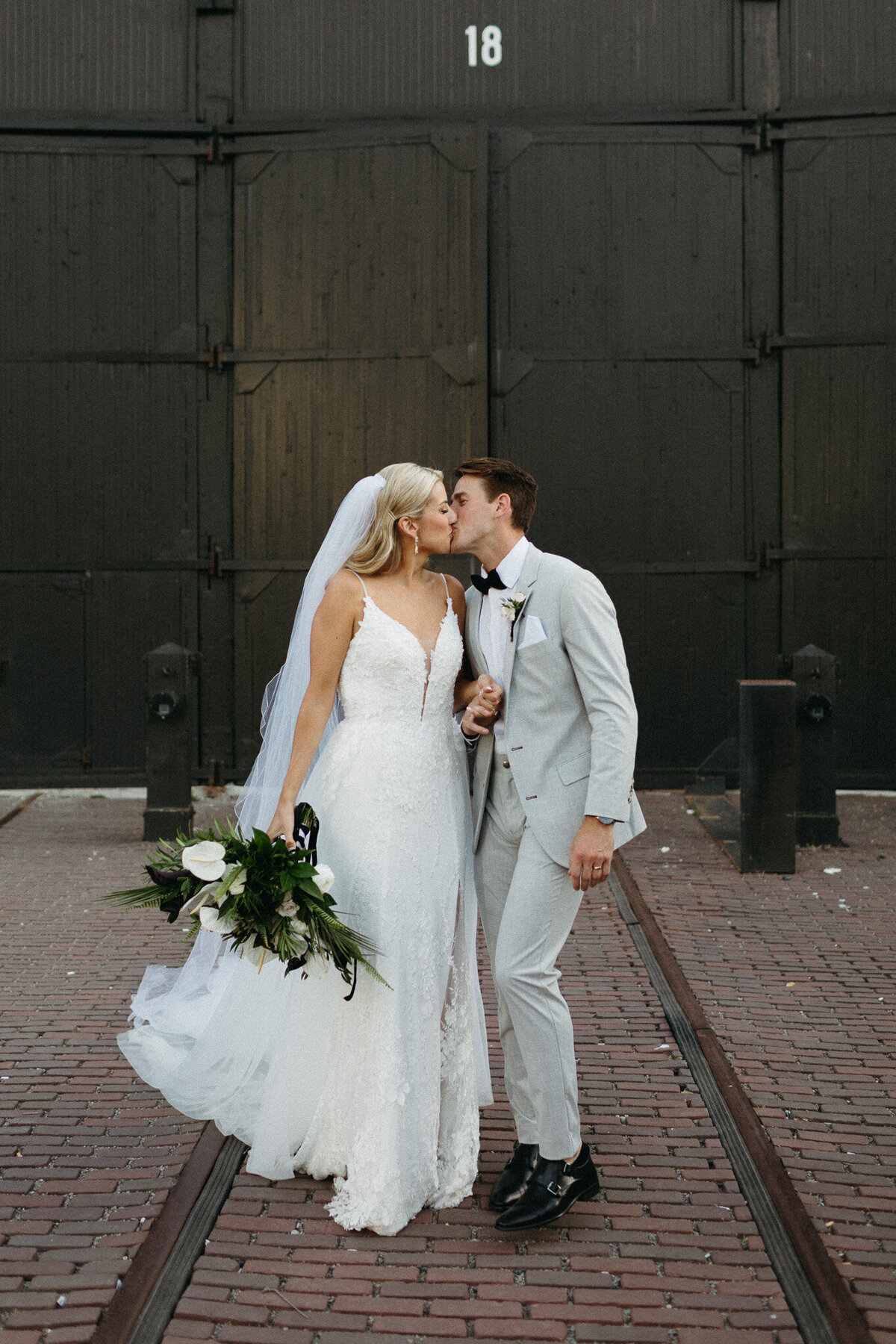 Bride in a white dress and veil holds a bouquet while kissing the groom in a light gray suit. They stand on brick pavement with black industrial doors behind them, captured beautifully at Steam Whistle Brewing by Toronto wedding photographer, Jennifer van Son.