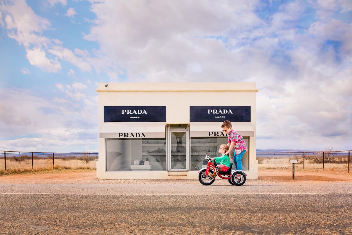 Two children pedldle a tricycle in front of the Prada Marfa art installation.
