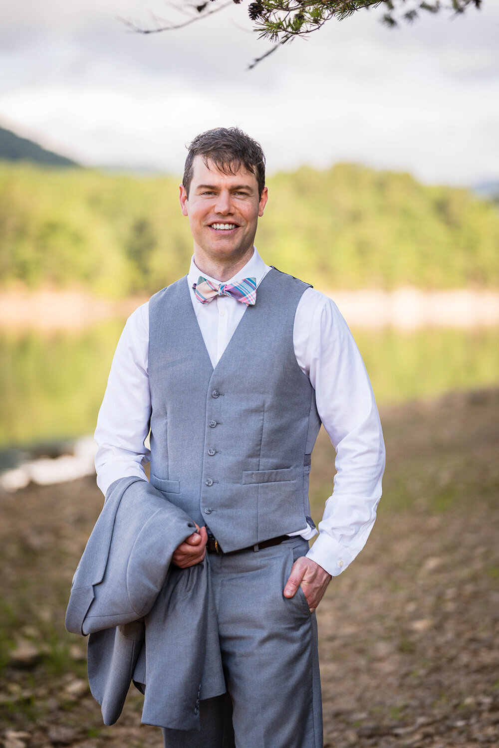 A groom stands along the shores of Carvin’s Cove for a formal portrait on his elopement day. He stands with one of his hands in his pocket and with his suit jacket draped over his other arm. He smiles and is wearing a plain white button up shirt, a pink and blue plaid bowtie, and gray vest and pants.