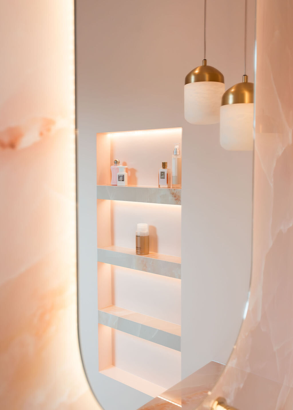 A minimalist bathroom shelf with built-in lighting contains various toiletries. Hanging above, two pendant lights with white and gold fixtures add a warm ambiance..