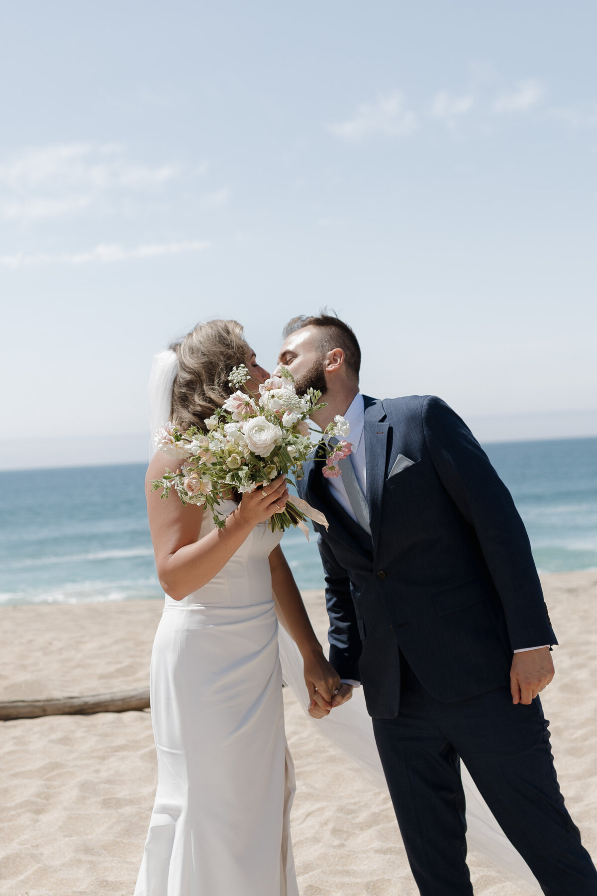 Bride and groom sharing a romantic kiss, holding bouquet on the sands of Montara State Beach near San Francisco.