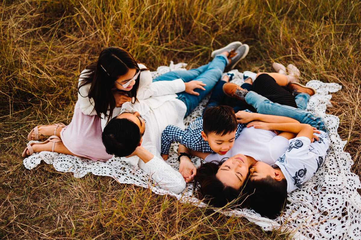 Family photo in the forest, they are lying on a white blanket and playing. The youngest child has a white T-shirt and is giving a kiss on the cheek to the woman. The man is looking up and is holding the older girl with glasses