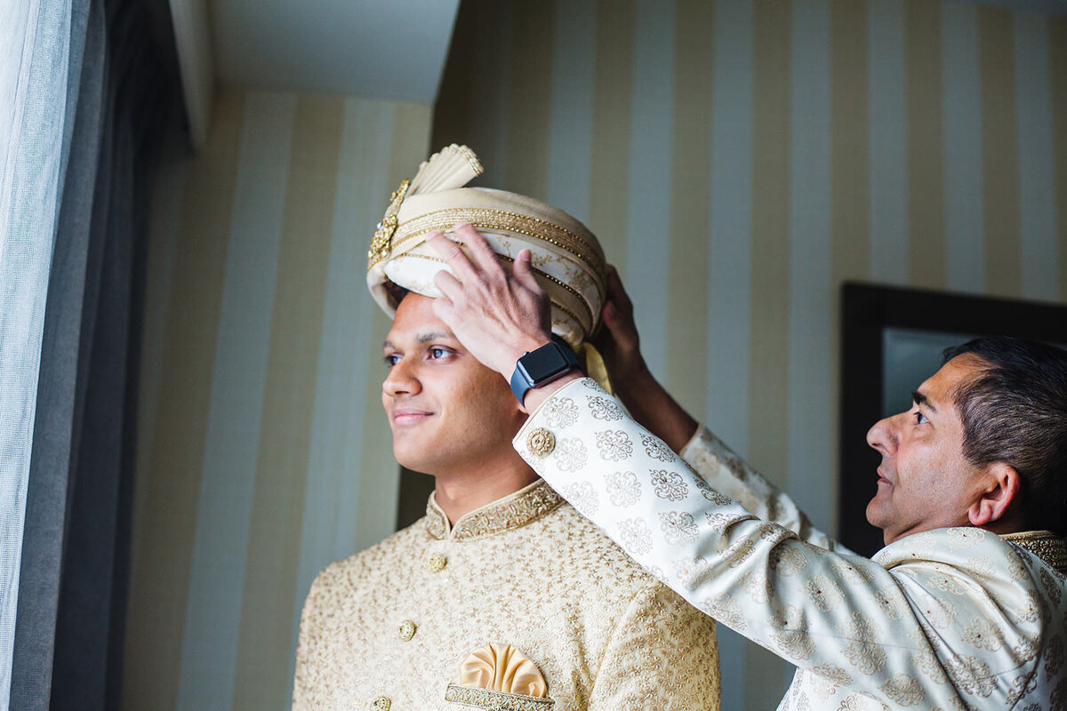A groom in an ornate cream sherwani and turban looks out the window while another man adjusts his headgear.