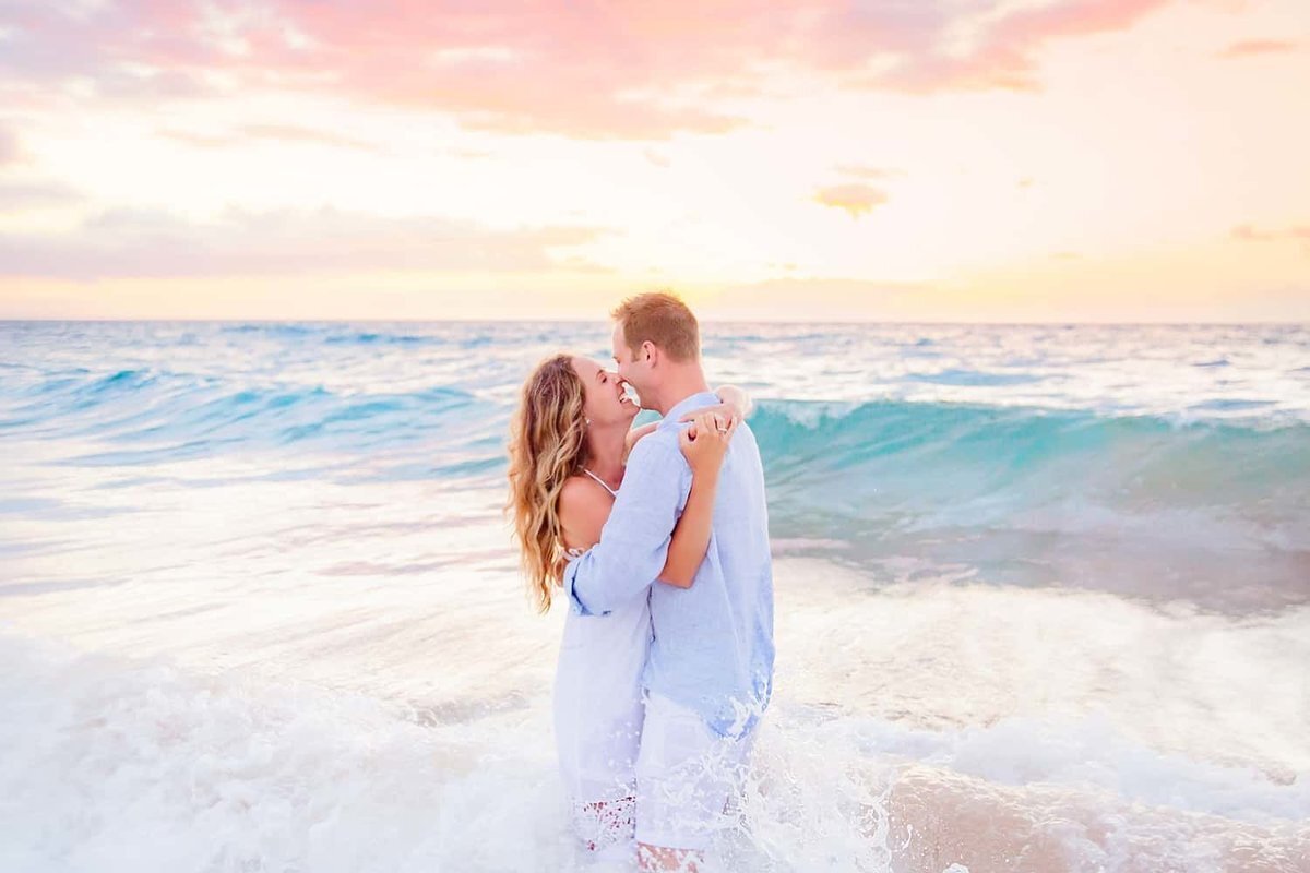 Beautiful couple kisses at sunset while honeymooning on Maui, waves crash on them as they hold one another under beautiful pastel skies