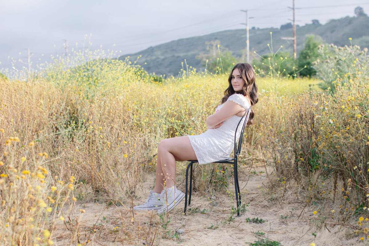 A person sits on a chair in a field of yellow wildflowers, wearing a white dress and gray sneakers. They are looking towards the camera with mountains in the background under a cloudy sky.