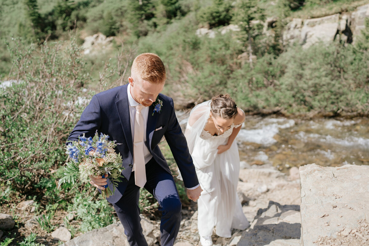 Groom hold bouquet and brides hand as helps her walk on the rocks near the waterfall  at their Ouray, Colorado Wedding