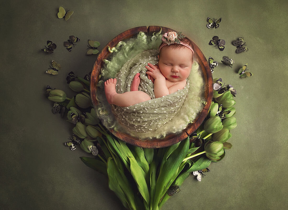 Snuggled in soft sage wrap, newborn sleeps in a wooden bowl surrounded by tulips and butterflies.