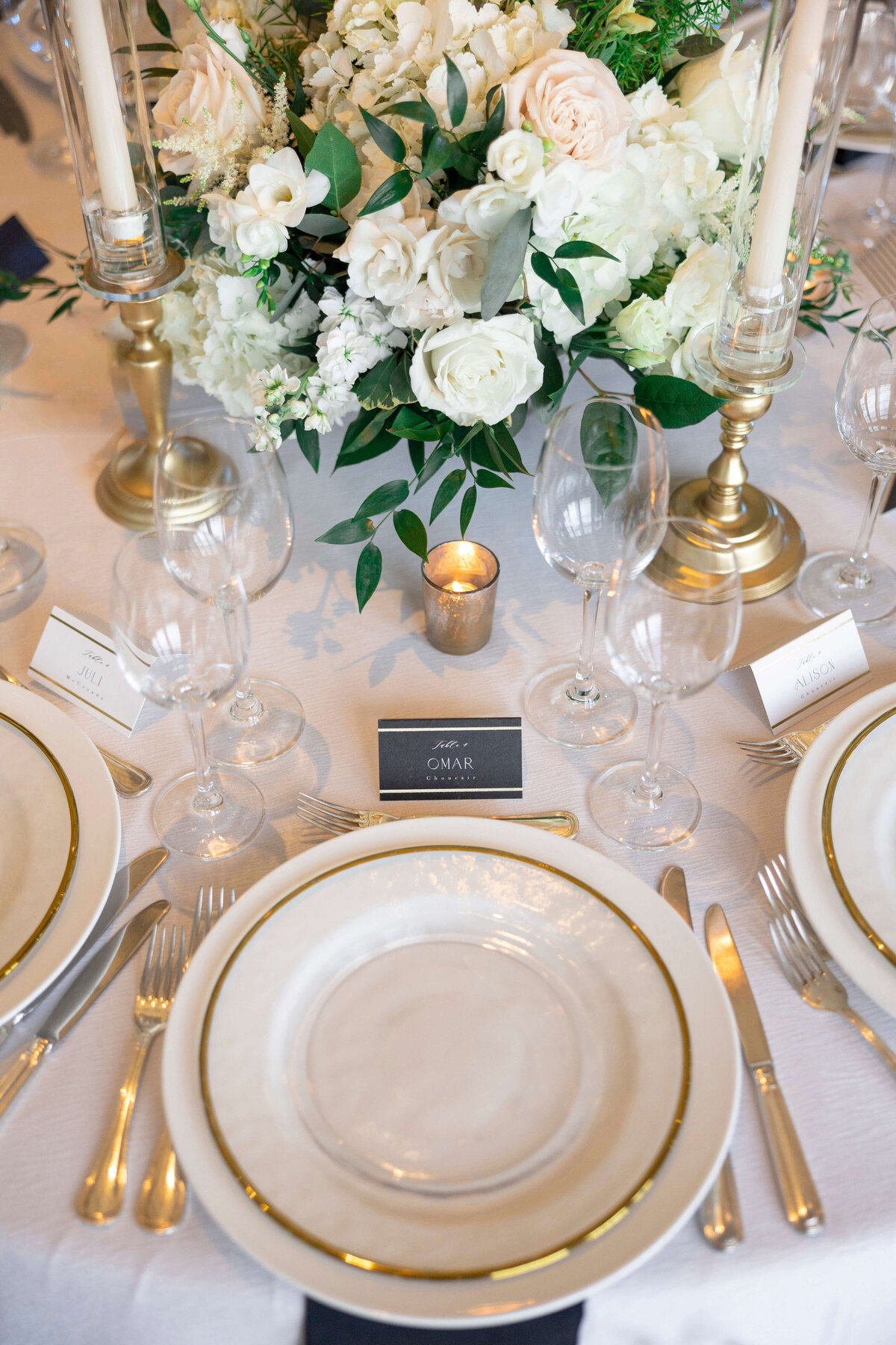 Elegant table setting with white plates, gold-rimmed edges, wine glasses, and cutlery. Centerpiece features white flowers and candles in gold holders.