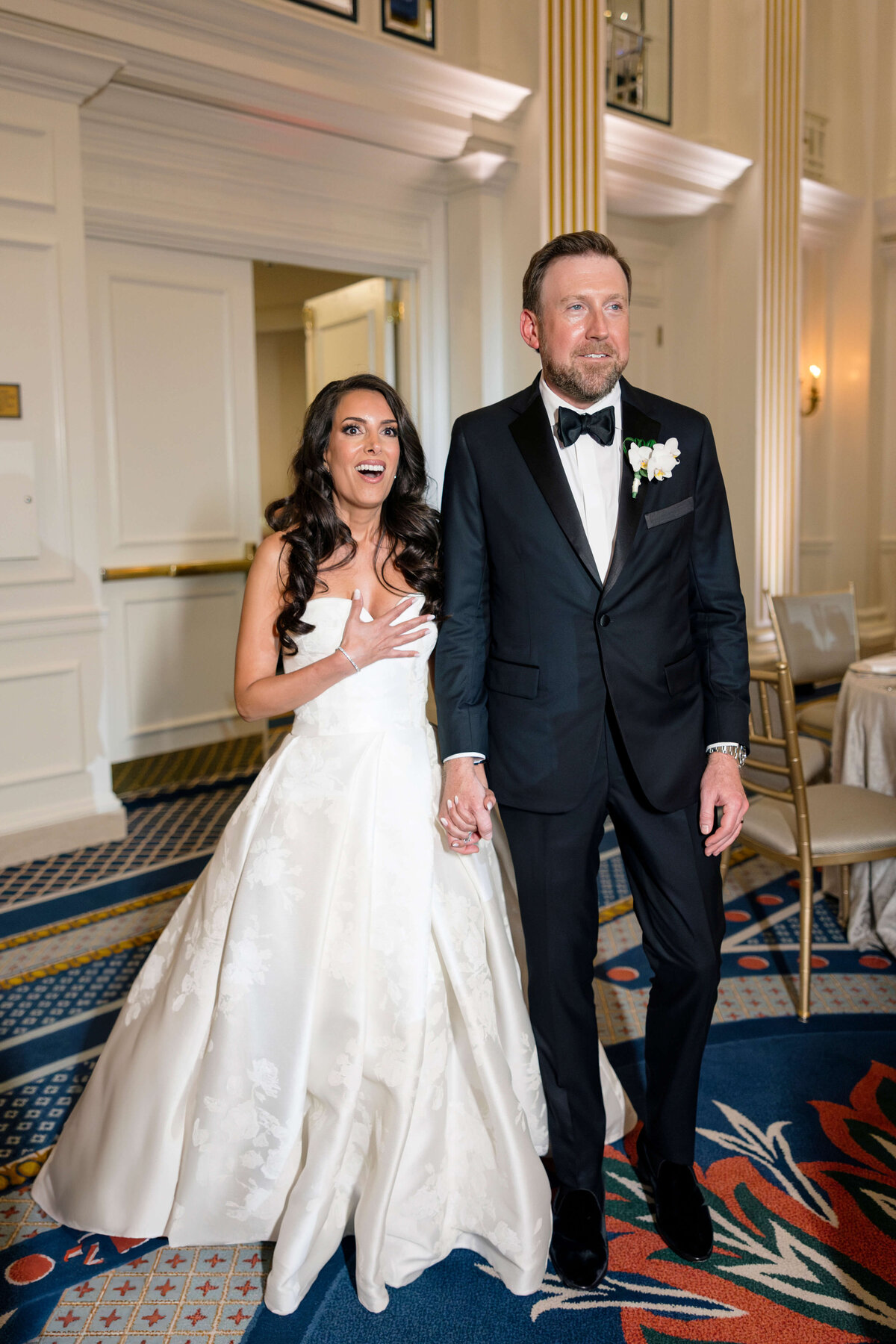 A bride and groom stand hand in hand in an elegant room. The bride wears a white gown, and the groom is in a black tuxedo with a bow tie. They both appear joyful and are looking in the same direction. The room features ornate decor and patterned carpeting.