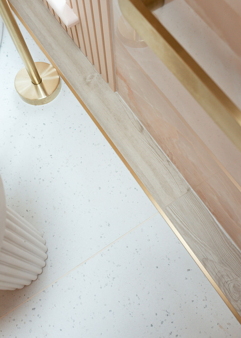 A close-up shows a section of a modern flooring. Light-colored speckled tiles are paired with wooden floor trim and brass accents.