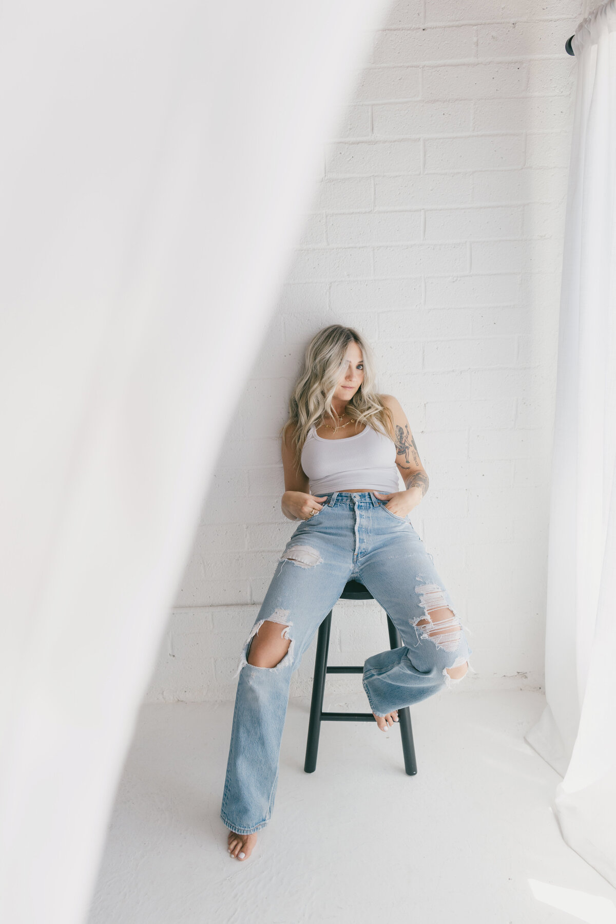 Minimal and editorial branding image of a woman in ripped jeans and a white tank sitting on a stool in a bright white studio — captured by Dani Lacey in Greenville SC.