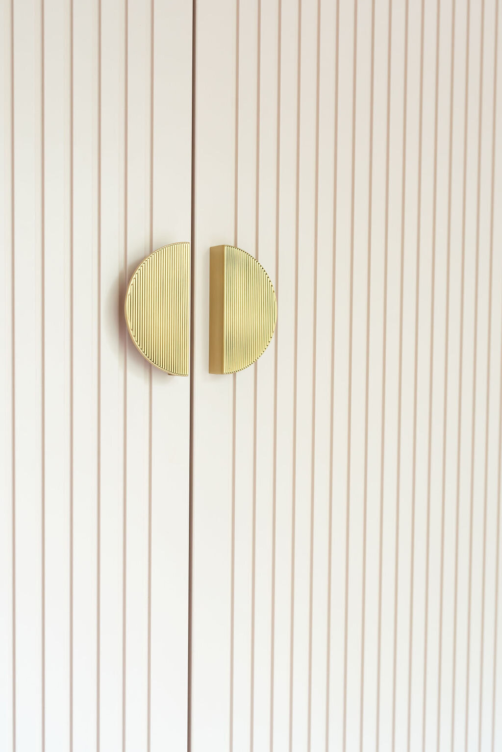 A close-up view of a modern  pale pink wardrobe with vertically striped doors and large circular gold handles.