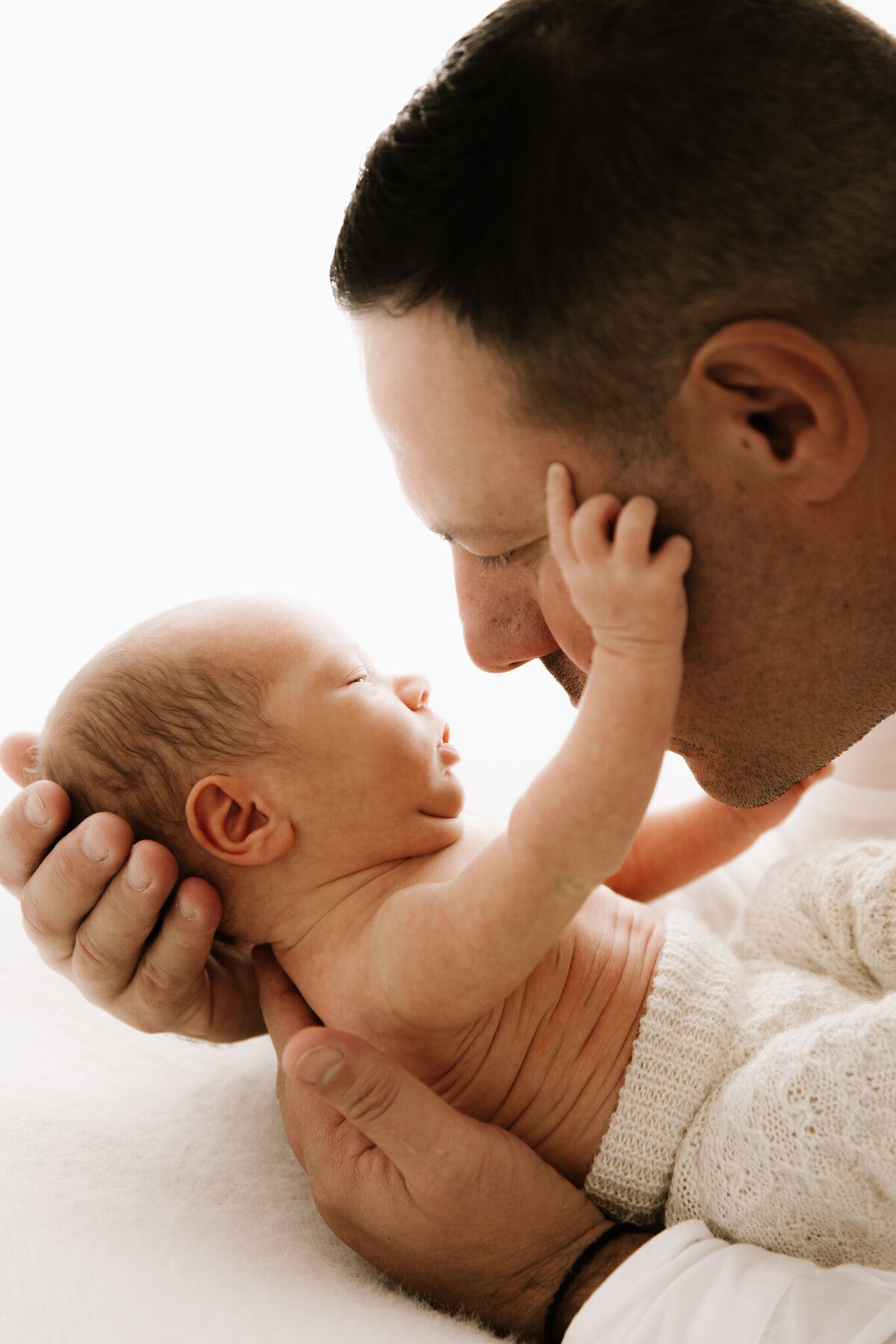 Newborn baby reaching up to touch their father’s face during a heartfelt moment captured in a softly lit photography studio in Southwest Sydney.